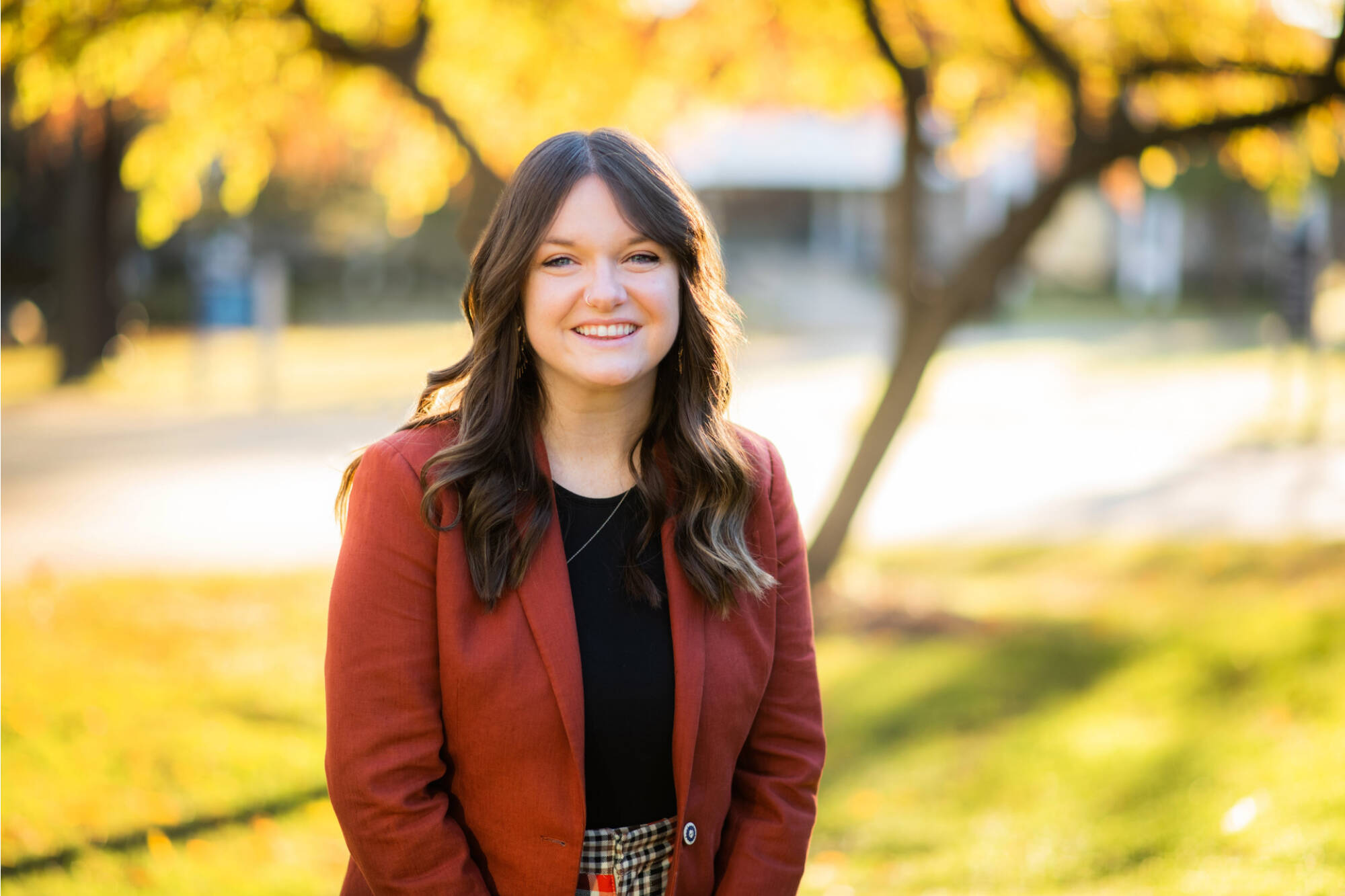 Picture of Em McKenna-Makela. Em is wearing a black shirt under a red suit coat. She has brown hair and is posed in front of a fall scene.